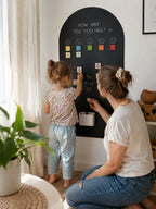 A woman kneels beside a young girl who is writing on a black arched wall chart in a bright room. The chart has smiley faces, color squares, and the words 'HOW ARE YOU FEELING?' and 'Be Kind'.
