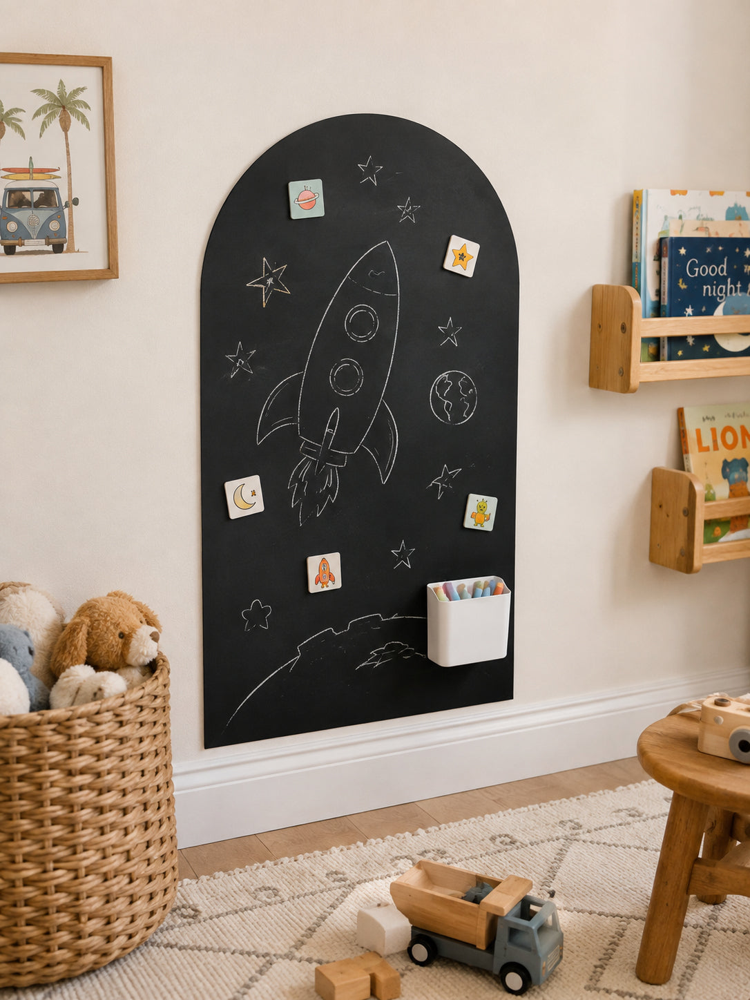 A black arched children's chalkboard with a white rocket, stars, and moon drawing, mounted on a beige wall in a styled children's room. The board has small magnetic cards and a white cup holder attached, with toys and book shelves visible around it.