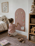A young child kneels on a textured rug in a nursery, writing on a dusty pink arch-shaped magnetic board. The board is decorated with small colorful stickers and a white cup holder.
