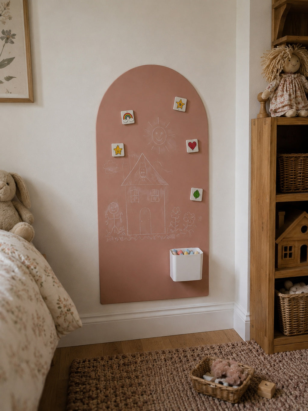 A dusty pink arched chalkboard wall hanging mounted on a cream wall in a child's bedroom. The board features a white chalk drawing of a house, small colorful magnetic tiles, and a white cup holder.
