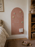 A dusty pink arched chalkboard wall hanging mounted on a cream wall in a child's bedroom. The board features a white chalk drawing of a house, small colorful magnetic tiles, and a white cup holder.
