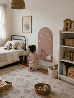 A young child kneels on a floral area rug in a styled children's bedroom, interacting with a dusty pink chalkboard-style wall decoration. The room features a bed, a white bookshelf with toys, and a woven rattan pendant light.