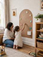 A woman and a young girl kneel on a wooden floor in a bright playroom, interacting with a large arch-shaped cork board wall. The girl is writing with a chalk-like stick, and small colorful cards are pinned to the board.