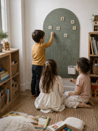 Three children are playing in a cozy, sunlit children's room. One child stands near a large arched chalkboard on the wall, writing and drawing, while the other two sit on a woven rug on the floor nearby.