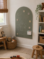 An arched sage green chalkboard wall hanging mounted on a white wall in a child's room. The board features hand-drawn white chalk illustrations of a house, sun, clouds, and trees, along with small colorful stickers and a white pencil cup.