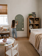 A young girl stands in a child's bedroom, writing on a green arched chalkboard mounted on the wall. The room features a floral-patterned bed, a wooden bookshelf, a small white table with chairs, and a window with a bamboo blind.