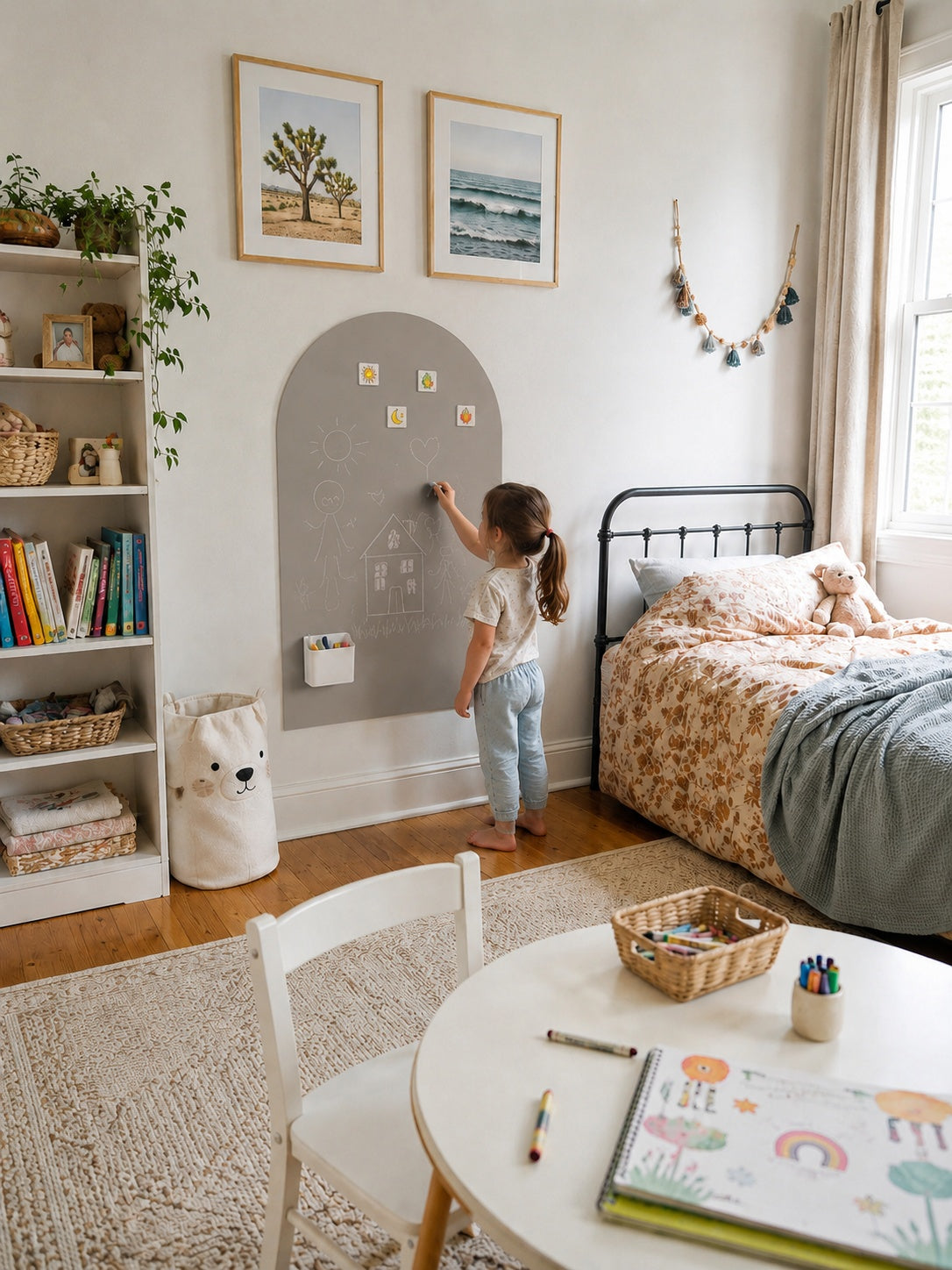 A child's bedroom scene with a young girl standing at a gray arch-shaped wall chalkboard, writing or drawing on it. The room includes a bed with floral bedding, a white bookshelf, a small round table with art supplies, and framed wall art.